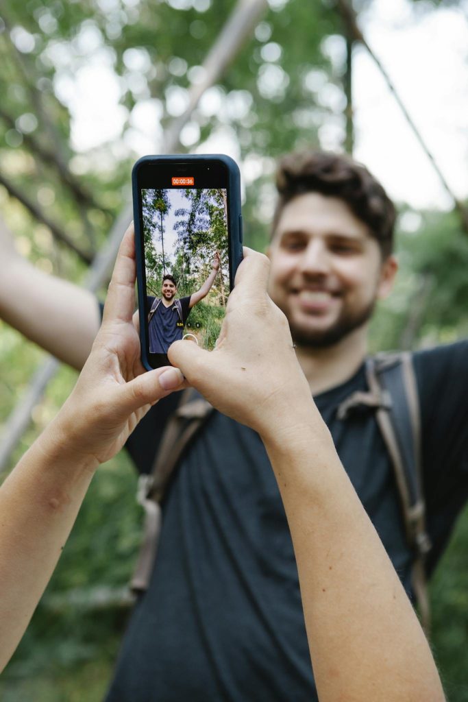 Crop unrecognizable woman taking photo of cheerful boyfriend on mobile phone while resting together in lush green jungle during summer vacation
