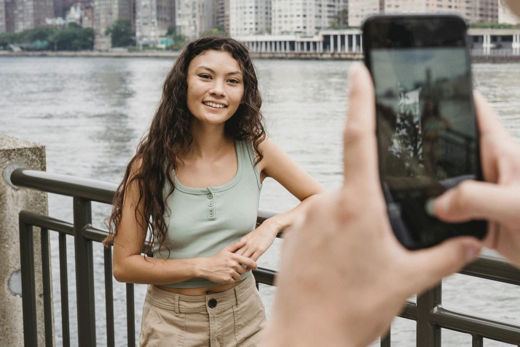 Unrecognizable person photographing smiling ethnic female friend on smartphone on embankment